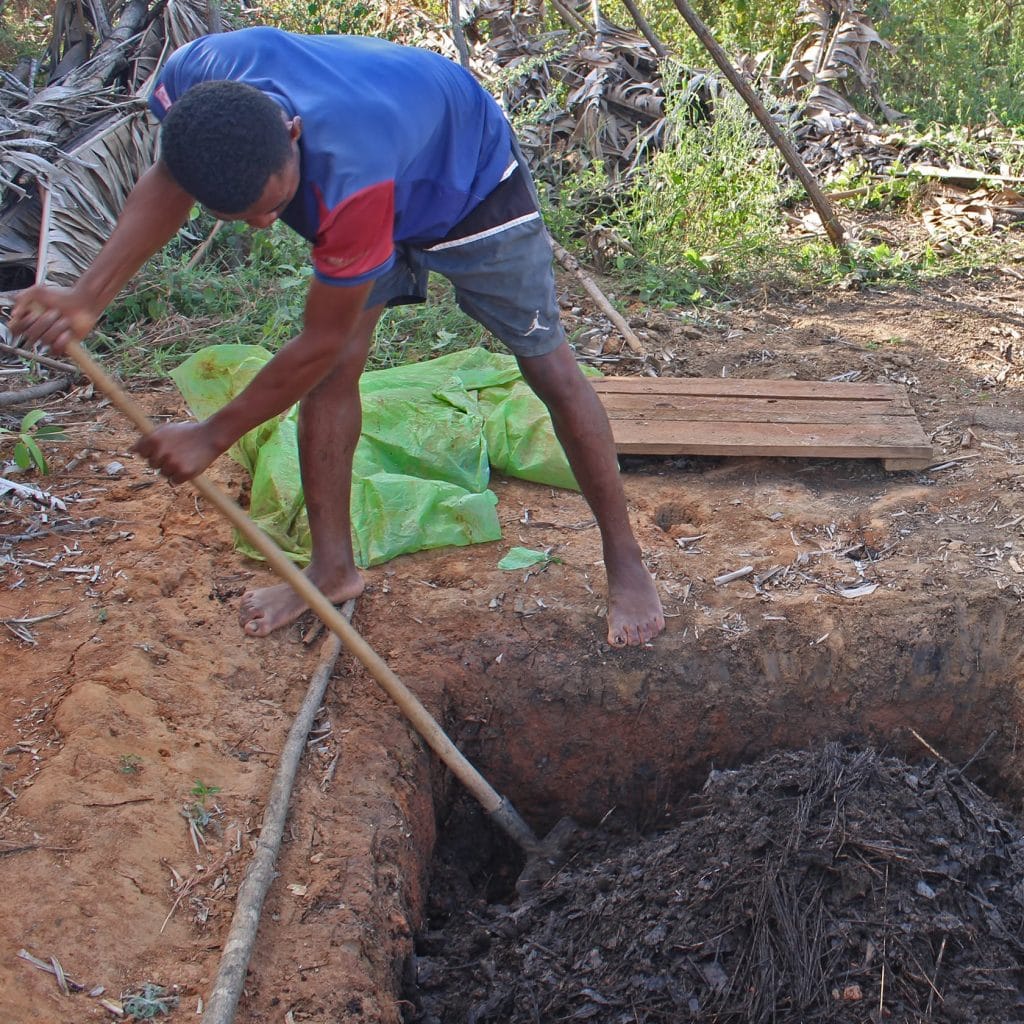 Man bending over, digging dirt with a shovel in a deep, wide hole in a dusty outdoor area; barefoot and wearing a blue shirt and shorts.