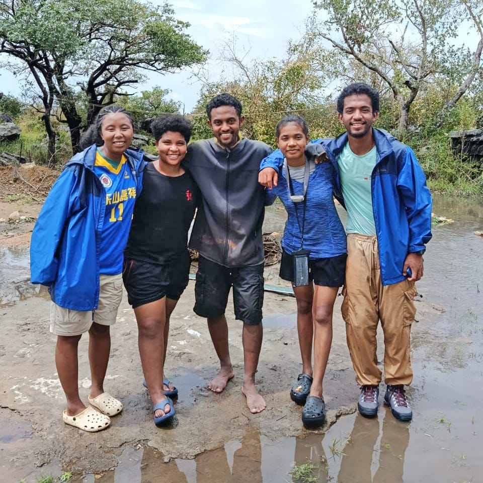 Five friends stand with arms around each other on a muddy outdoor path, smiling in casual clothes and jackets after rain.