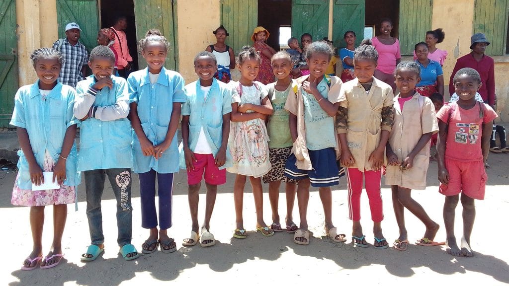 Group of children in blue uniforms posing outside a building with green doors, smiling at the camera.
