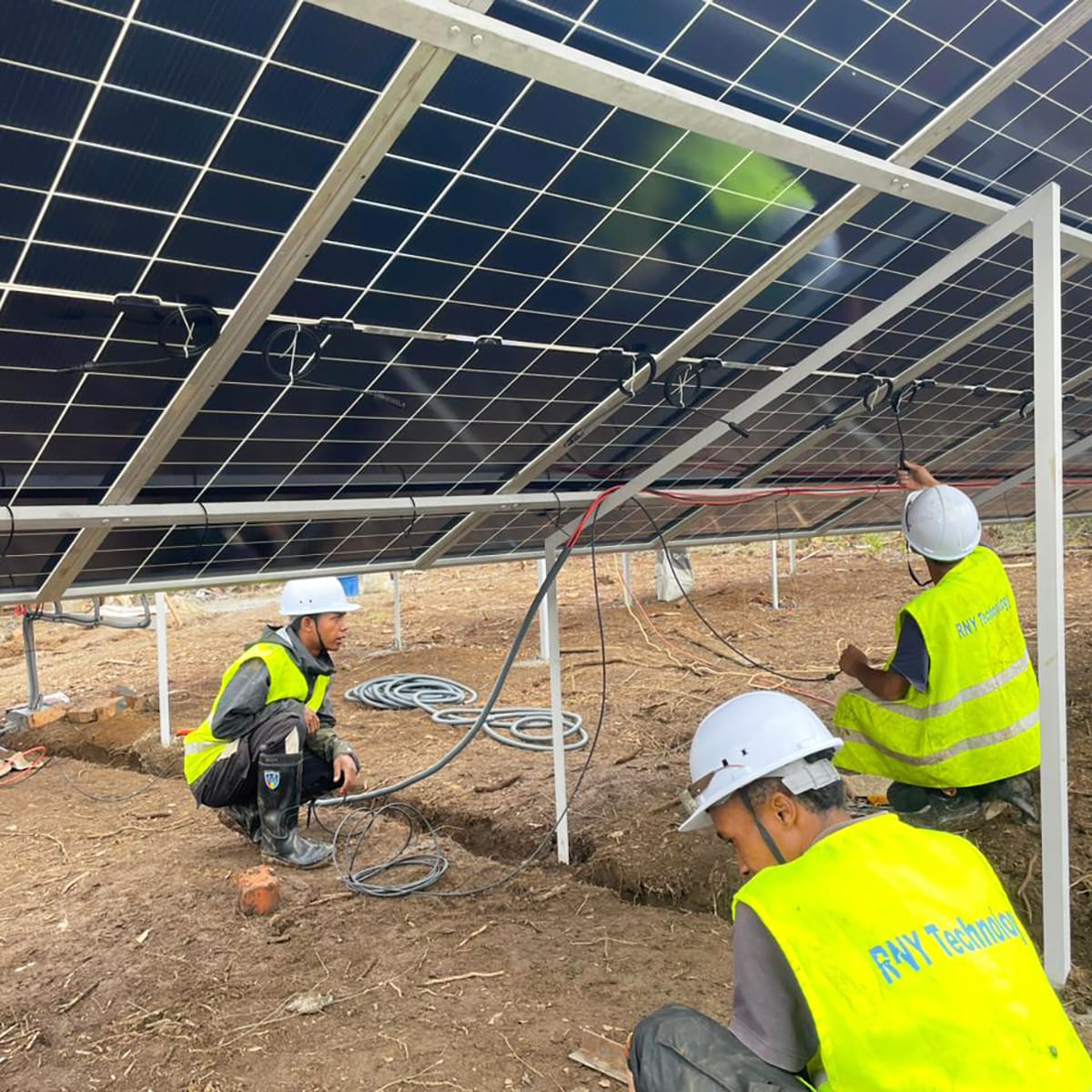 Construction workers in safety vests and helmets installing wiring under solar panels in a solar farm on a dirt site.