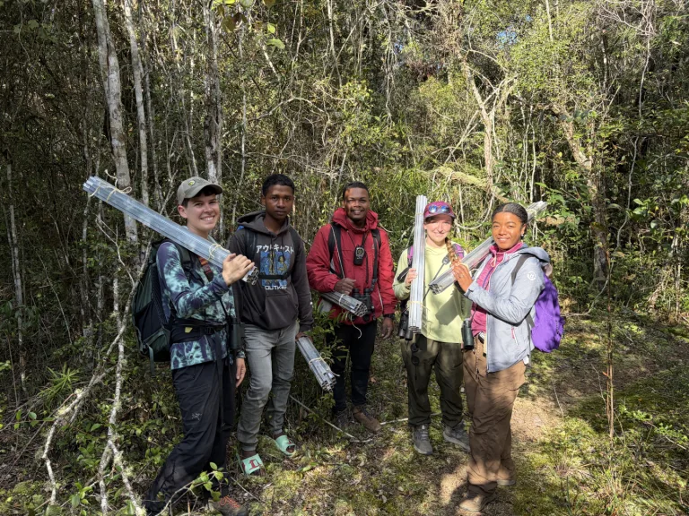 Group of five hikers in a forest, each holding bundles of metal tubing and wearing backpacks while posing for a photo.