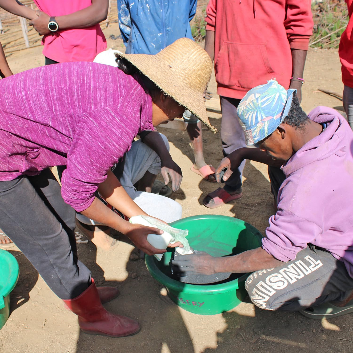 People washing items in a green basin outdoors, kneeling in the dirt while others stand nearby; one person wears a straw hat and a purple hoodie.