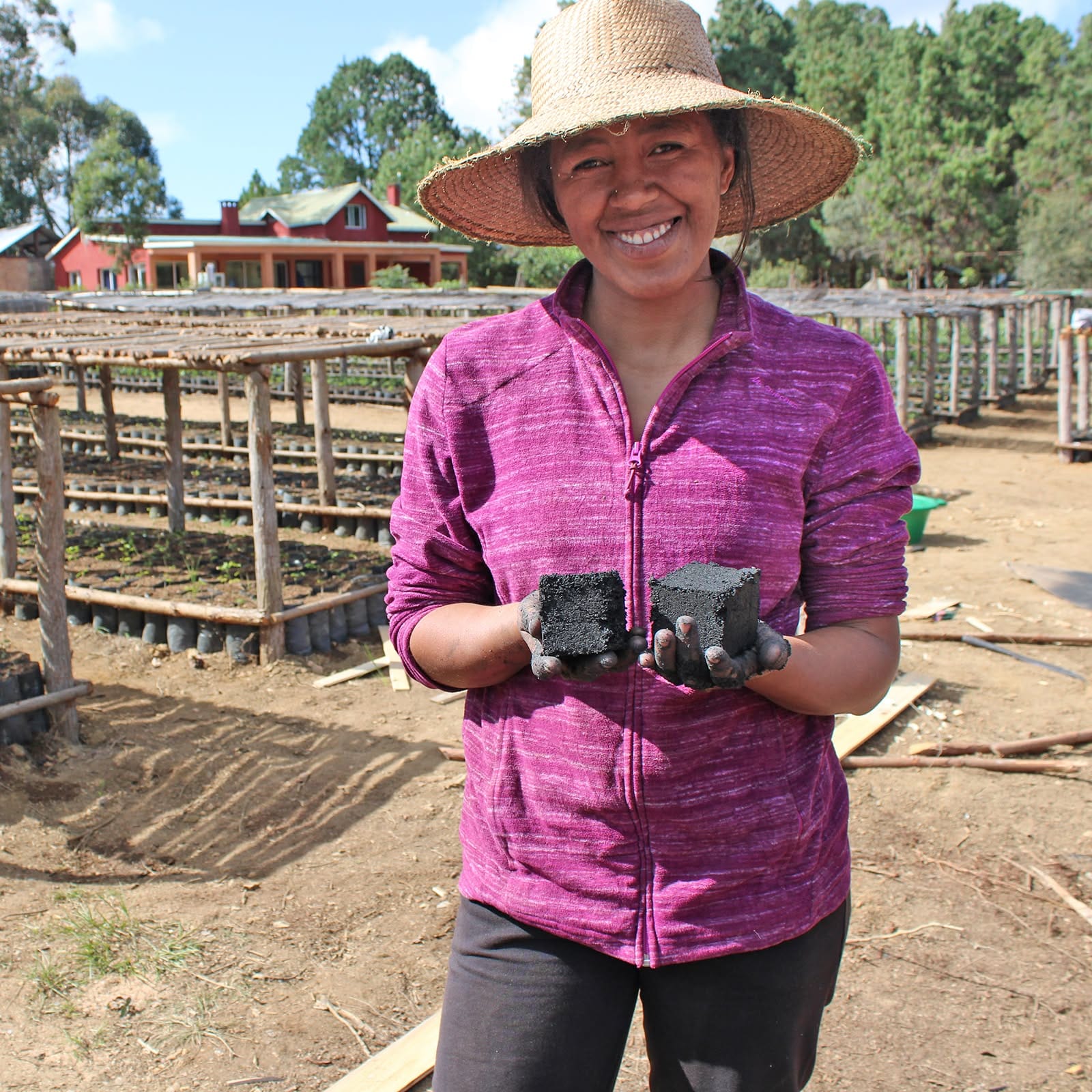 Smiling woman in a wide straw hat and magenta jacket holds two dark blocks in her hands at a farm site.