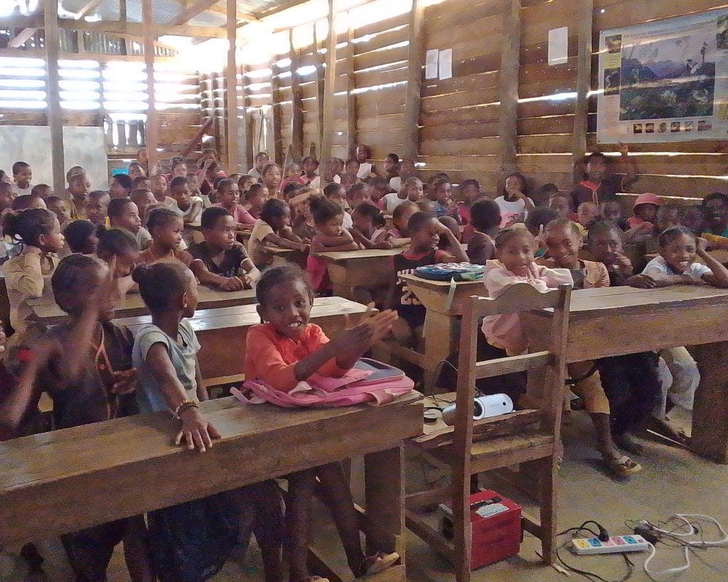 Group of schoolchildren seated at wooden benches in a bright, rustic classroom.