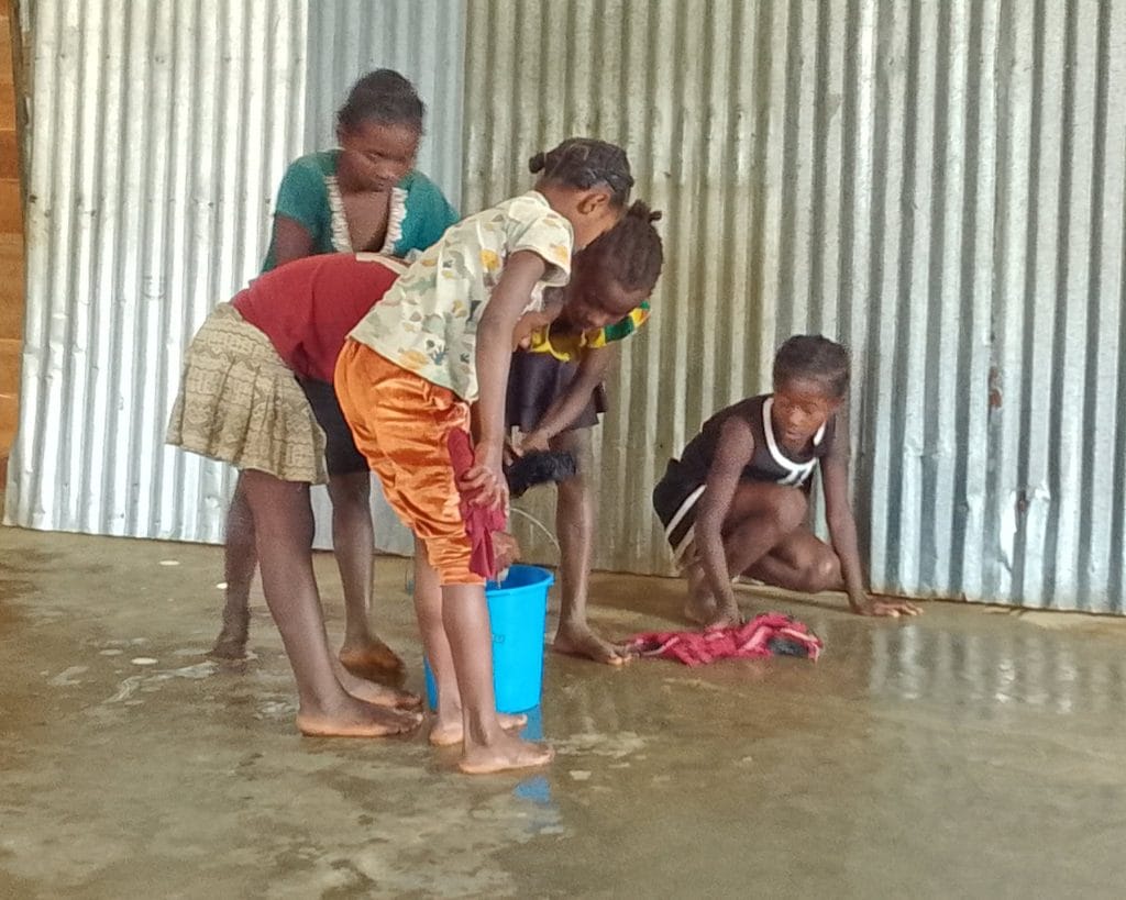 Children washing clothes in a blue bucket on a wet concrete floor near a corrugated metal wall, barefoot and bent over.