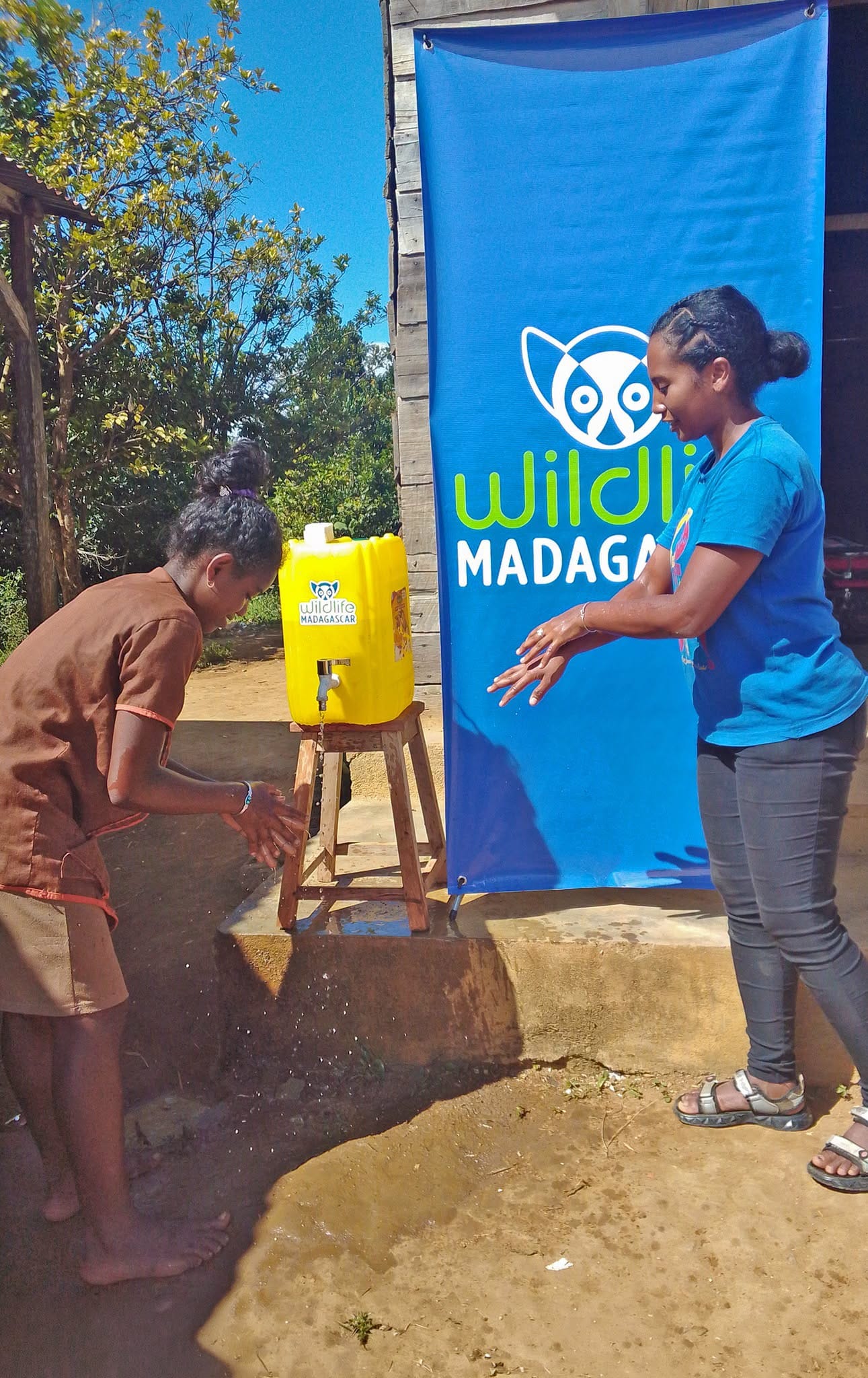 Two people wash their hands at an outdoor handwashing station next to a yellow water container labeled Wildlife Madagascar, with a blue Willdlife Madagascar banner in the background.