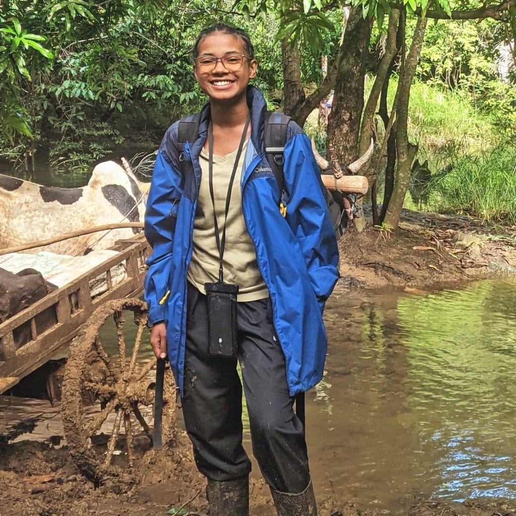 Smiling person in a blue rain jacket standing in a muddy riverside trail with a backpack and glasses.