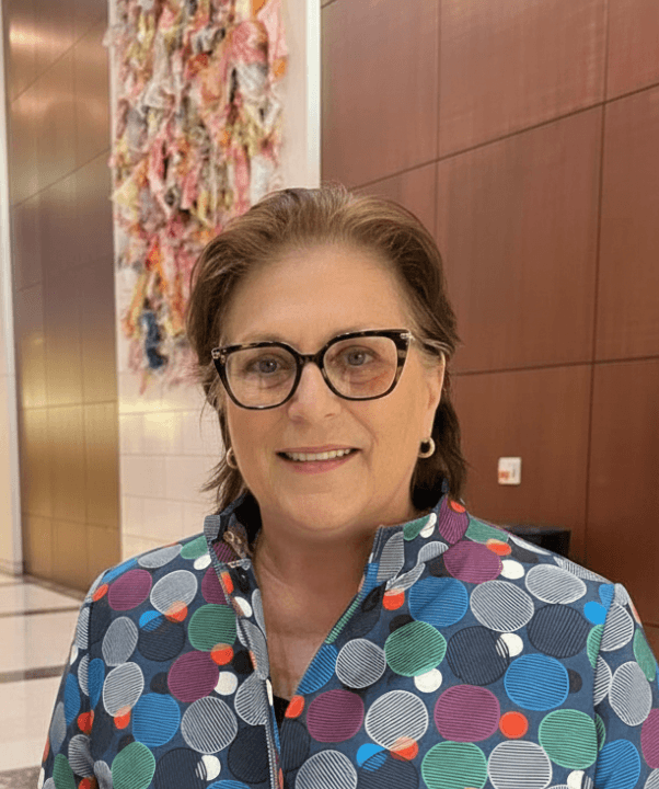 Smiling woman with short brown hair and black-framed glasses wearing a colorful polka-dot jacket in a modern lobby with abstract wall art behind her.
