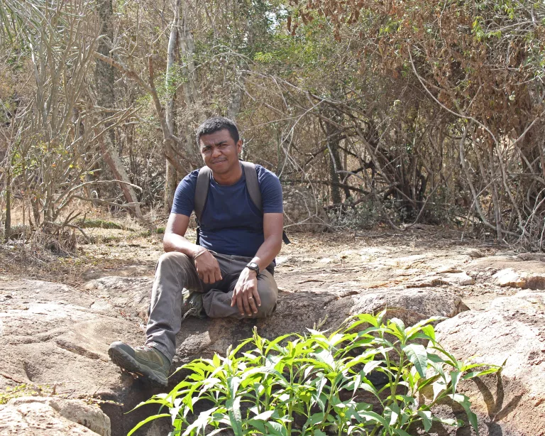 Man sits on rocky terrain outdoors in dry, scrubby woodland, wearing a blue shirt and backpack, looking toward the camera.