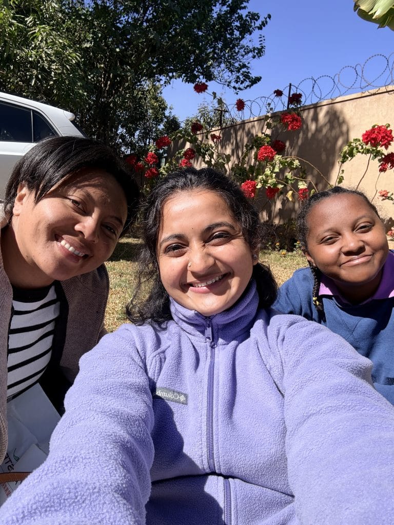 Three women pose for a sunny outdoor selfie with red flowers and a wall behind them.