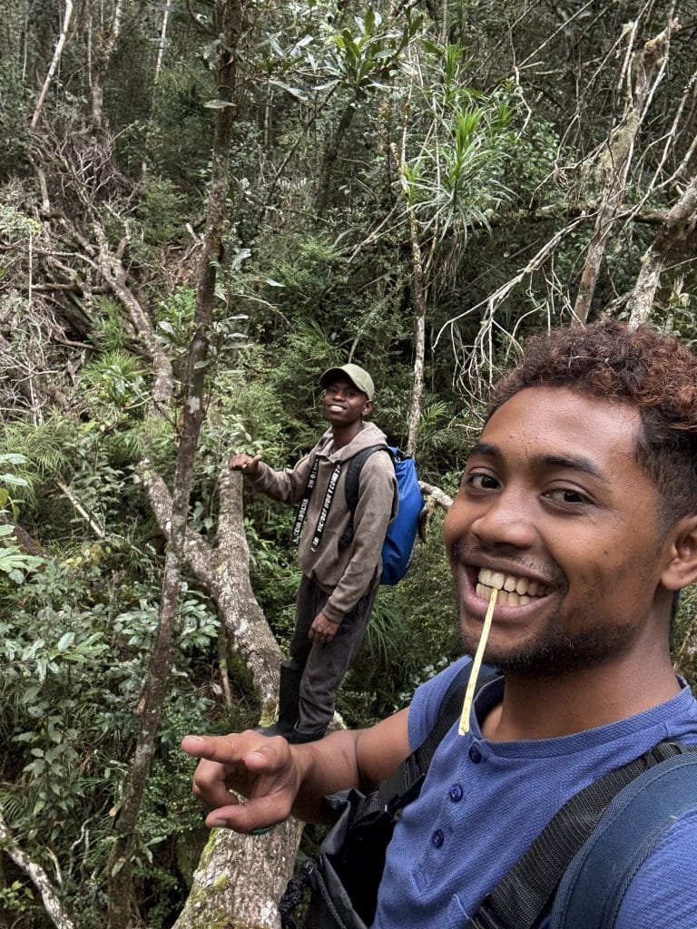 Two hikers in a dense forest; foreground man smiles at the camera with a toothpick, wearing a blue shirt and a backpack, with another hiker in a cap in the background.