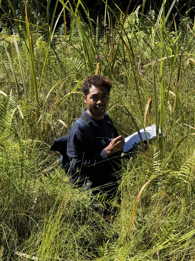 Man standing in tall grass, holding a notebook and pen, looking toward the camera with a backpack strap visible