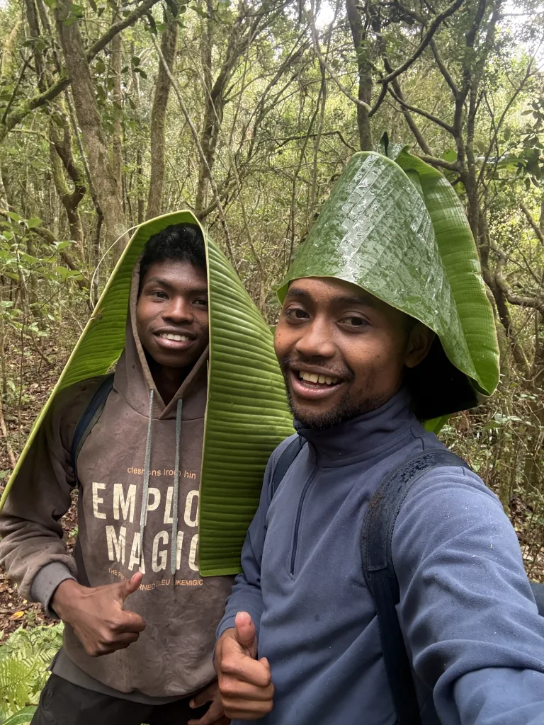 Two men pose for a selfie in a dense forest, wearing oversized banana leaf hats and smiling at the camera.