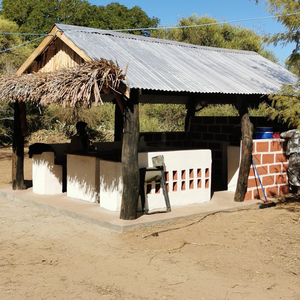 Open shelter with a metal roof, palm frond pile on the left, white counter, and a person seated in the shade under the structure.