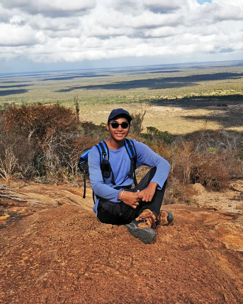 Hiker wearing a blue long-sleeve shirt and backpack sits on rocky red ground with a vast desert plain and cloudy sky behind.