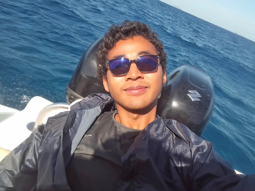 Young man wearing sunglasses and a dark jacket taking a selfie on a boat with the ocean in the background.
