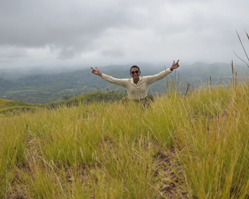 Man standing in tall grass with arms raised in a hilly, cloudy landscape.
